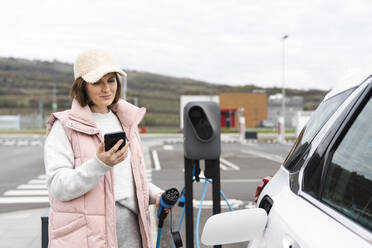 Woman holding smart phone and electric charger by car at station - EKGF00194