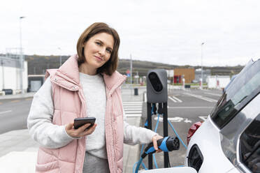 Frau mit Ladegerät und Smartphone am Elektroauto an der Tankstelle - EKGF00196