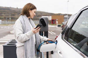 Frau mit Smartphone lädt Elektroauto an der Tankstelle - EKGF00200