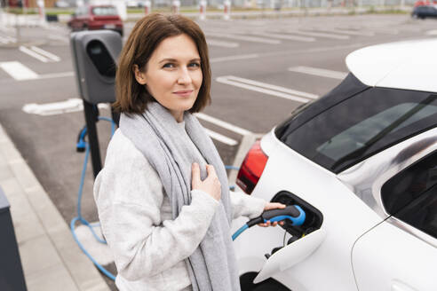 Lächelnde Frau beim Aufladen eines Elektroautos an einer Tankstelle - EKGF00202