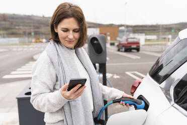 Woman using smart phone charging electric car at station - EKGF00204