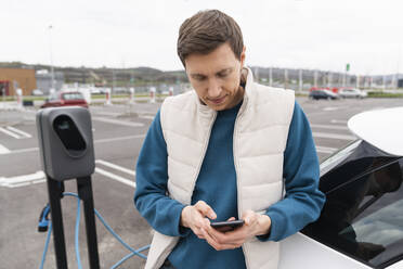 Mann mit Smartphone, der sich an einer Ladestation an ein Auto lehnt - EKGF00205