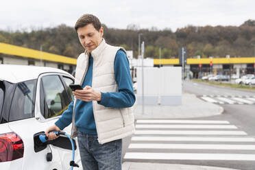 Mann mit Smartphone neben einem Elektroauto am Bahnhof - EKGF00217