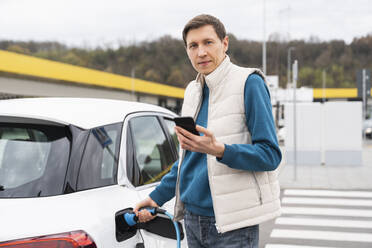 Mann, der sein Auto auflädt und dabei sein Smartphone in der Hand hält - EKGF00218