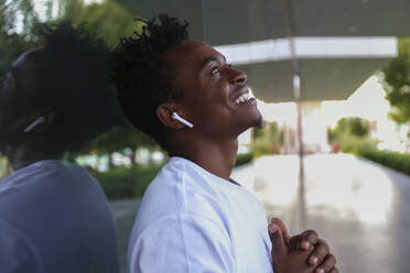 Lächelnder Mann mit kabellosen In-Ear-Kopfhörern, der an der Wand lehnt und träumt - SYEF00030