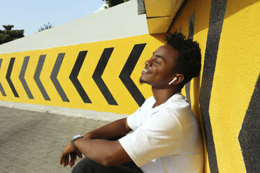 Smiling man with eyes closed listening to music sitting near wall - SYEF00038