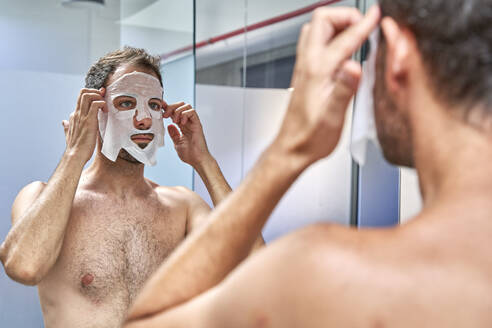 Man applying sheet mask on face in front of mirror - VEGF06098