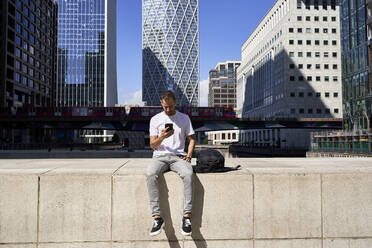 Mann mit Smartphone vor einem Gebäude an der Wand sitzend - VEGF06116