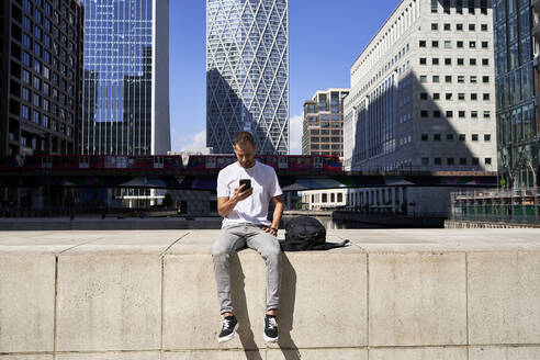 Man using smart phone sitting on wall in front of buildings - VEGF06116