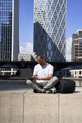 Man using laptop in front of buildings on sunny day - VEGF06119