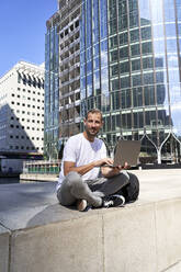 Smiling man with laptop sitting in front of buildings - VEGF06121