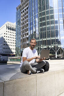 Lächelnder Mann mit Laptop vor einem Gebäude sitzend - VEGF06121