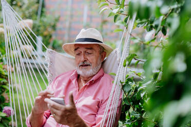 Glücklicher älterer Mann sitzt auf einer Gartenschaukel und scrollt auf seinem Smartphone. - HPIF03710