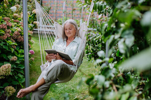 Ältere Frau entspannt sich auf einer Gartenschaukel mit einem Buch. - HPIF03714