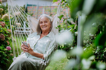 Ältere Frau entspannt sich auf einer Gartenschaukel mit einem Smartphone. - HPIF03715