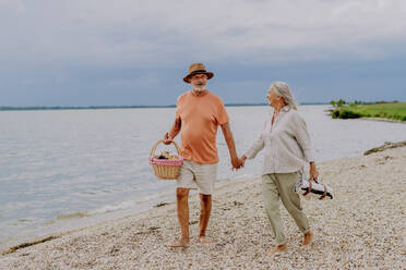 Älteres Paar beim Spaziergang mit Picknickkorb am Meer. - HPIF03743