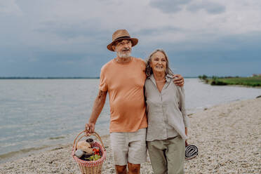 Älteres Paar beim Spaziergang mit Picknickkorb am Meer. - HPIF03744