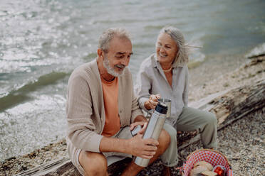 Ein älteres Ehepaar sitzt in der Nähe des herbstlichen Flusses, ruht sich aus und trinkt Tee aus einer Thermoskanne. - HPIF03753
