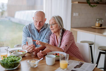 Ein glückliches älteres Ehepaar benutzt sein Smartphone beim gemeinsamen Abendessen zu Hause. - HPIF03879