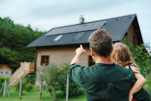 Rear view of dad holding her little girl in arms and showing at their house with solar panels.Alternative energy, saving resources and sustainable lifestyle concept. - HPIF03953