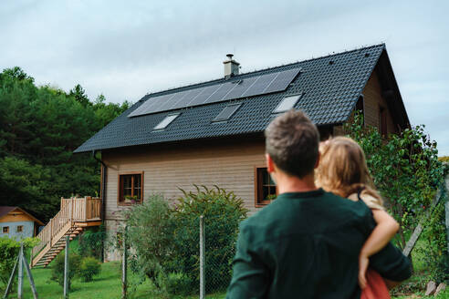 Rückansicht des Vaters, der sein kleines Mädchen in den Armen hält und auf sein Haus mit Sonnenkollektoren blickt: Alternative Energie, Ressourceneinsparung und nachhaltiger Lebensstil. - HPIF03954