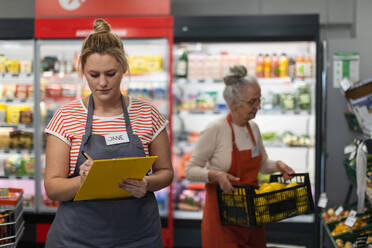 Eine junge Verkäuferin im Supermarkt in der Gemüseschale, im Hintergrund ihr Kollege beim Auffüllen der Vorräte. - HPIF04000