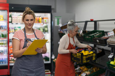 Eine junge Verkäuferin im Supermarkt in der Gemüseschale, im Hintergrund ihr Kollege beim Auffüllen der Vorräte. - HPIF04002