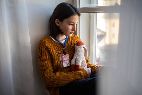 A sad Ukrainian refugee schoolgirl missing home and looking through window. Ukrainian war concept. - HPIF04064