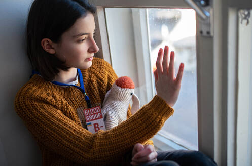 A sad Ukrainian refugee schoolgirl missing home and looking through window. Ukrainian war concept. - HPIF04065