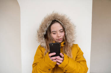 Eine glückliche junge Frau mit Down-Syndrom, die einen Parka trägt, sich an die Wand lehnt und ihr Smartphone benutzt. - HPIF04079
