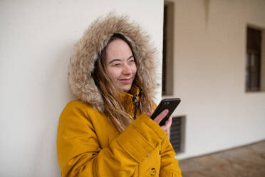 Eine glückliche junge Frau mit Down-Syndrom, die einen Parka trägt, sich an die Wand lehnt und ihr Smartphone benutzt. - HPIF04080