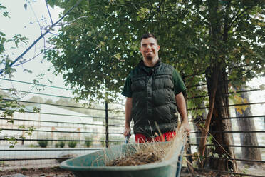 Caretaker with down syndrome in the zoo pushing wheelbarrow after tidying animal enclosure. Concept of integration people with disabilities into society. - HPIF04092