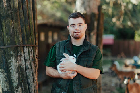 Caretaker with down syndrome taking care of animals in zoo, stroking a rabbit. Concept of integration people with disabilities into society. - HPIF04099