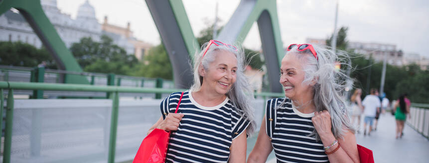 Glückliche ältere Zwillinge in gleicher Kleidung, die in einer Stadt spazieren gehen und vom Einkaufen zurückkehren. - HPIF04183