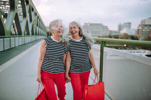 Glückliche ältere Zwillinge in gleicher Kleidung, die in einer Stadt spazieren gehen und vom Einkaufen zurückkehren. - HPIF04189