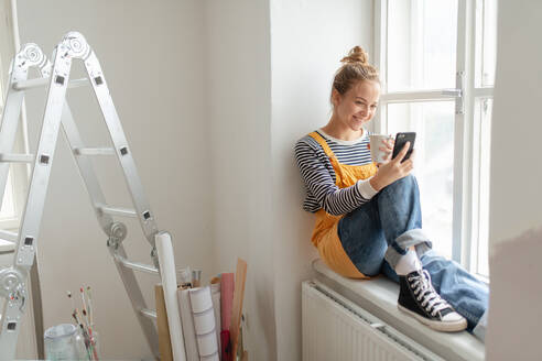 Glückliche junge Frau, die während der Renovierung ihres Hauses eine Pause einlegt, telefoniert und in den sozialen Medien nach Inspiration sucht. - HPIF04203