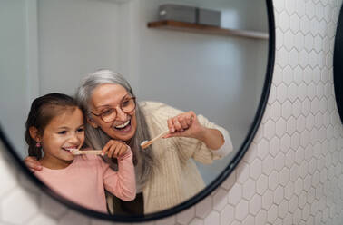 A grandmother with granddaughter standing indoors in bathroom, brusing teeth and looking at mirror. - HPIF04211