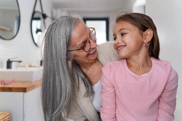 A senior grandmother and granddaughter standing indoors in bathroom, looking at each other. - HPIF04218