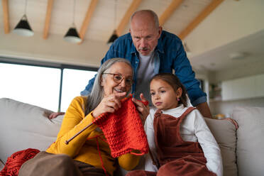 A little girl sitting on sofa with her grandparents and learning to knit indoors at home. - HPIF04225
