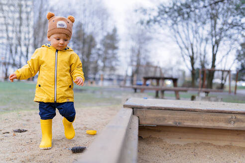 Fröhlicher kleiner Junge mit einer Teddybär-Mütze, der auf einem Spielplatz im Sandkasten spielt, während eines Herbsttages. - HPIF04266
