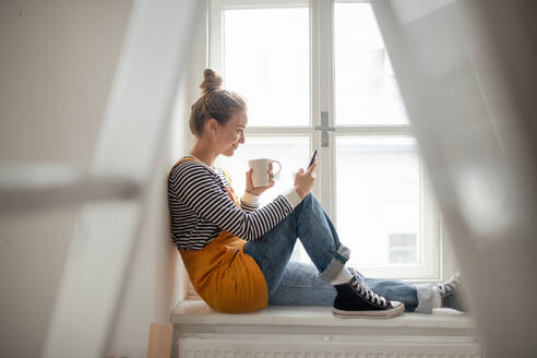 Glückliche junge Frau, die während der Renovierung ihres Hauses eine Pause einlegt, telefoniert und in den sozialen Medien nach Inspiration sucht. - HPIF04277