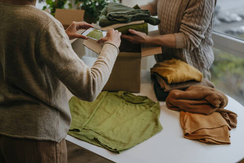 Junge Modedesignerin, die mit ihrem Smartphone fotografiert, während ein Kollege Kleidung in einen Karton packt - YTF00382