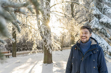 Glücklicher Mann in der Nähe eines verschneiten Baumes im Park - ANAF00672