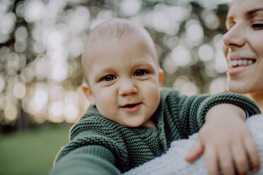 Eine Mutter hält ihren kleinen Sohn in einem gestrickten Pullover während eines Spaziergangs in der Natur und schaut in die Kamera. - HPIF04401