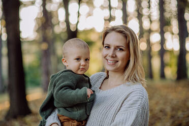 Eine Mutter hält ihren kleinen Sohn in einem gestrickten Pullover während eines Spaziergangs in der Natur und schaut in die Kamera. - HPIF04402