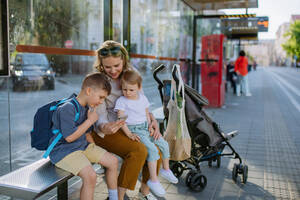 Eine junge Mutter mit kleinen Kindern wartet an einer Bushaltestelle in der Stadt und scrollt auf ihrem Mobiltelefon. - HPIF04452