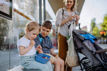 Eine junge Mutter mit kleinen Kindern wartet an einer Bushaltestelle in der Stadt und isst einen Obstsnack. - HPIF04457