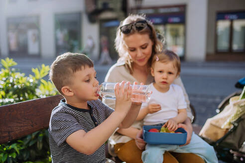 Eine junge Mutter mit kleinen Kindern sitzt im Sommer auf einer Bank in der Stadt, isst einen Obstsnack und trinkt Wasser. - HPIF04474