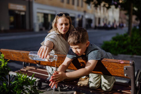 Eine junge Mutter hält eine abfallfreie Wasserflasche in der Hand und wäscht ihrem kleinen Sohn die Hände, während sie auf einer Bank in der Stadt sitzt, nachhaltiger Lebensstil. - HPIF04475