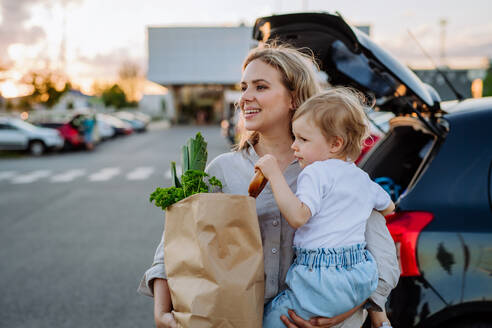 Eine junge Mutter mit ihrer kleinen Tochter hält nach dem Einkaufen abfallfreie Einkaufstüten mit Lebensmitteln in der Nähe des Autos. - HPIF04483